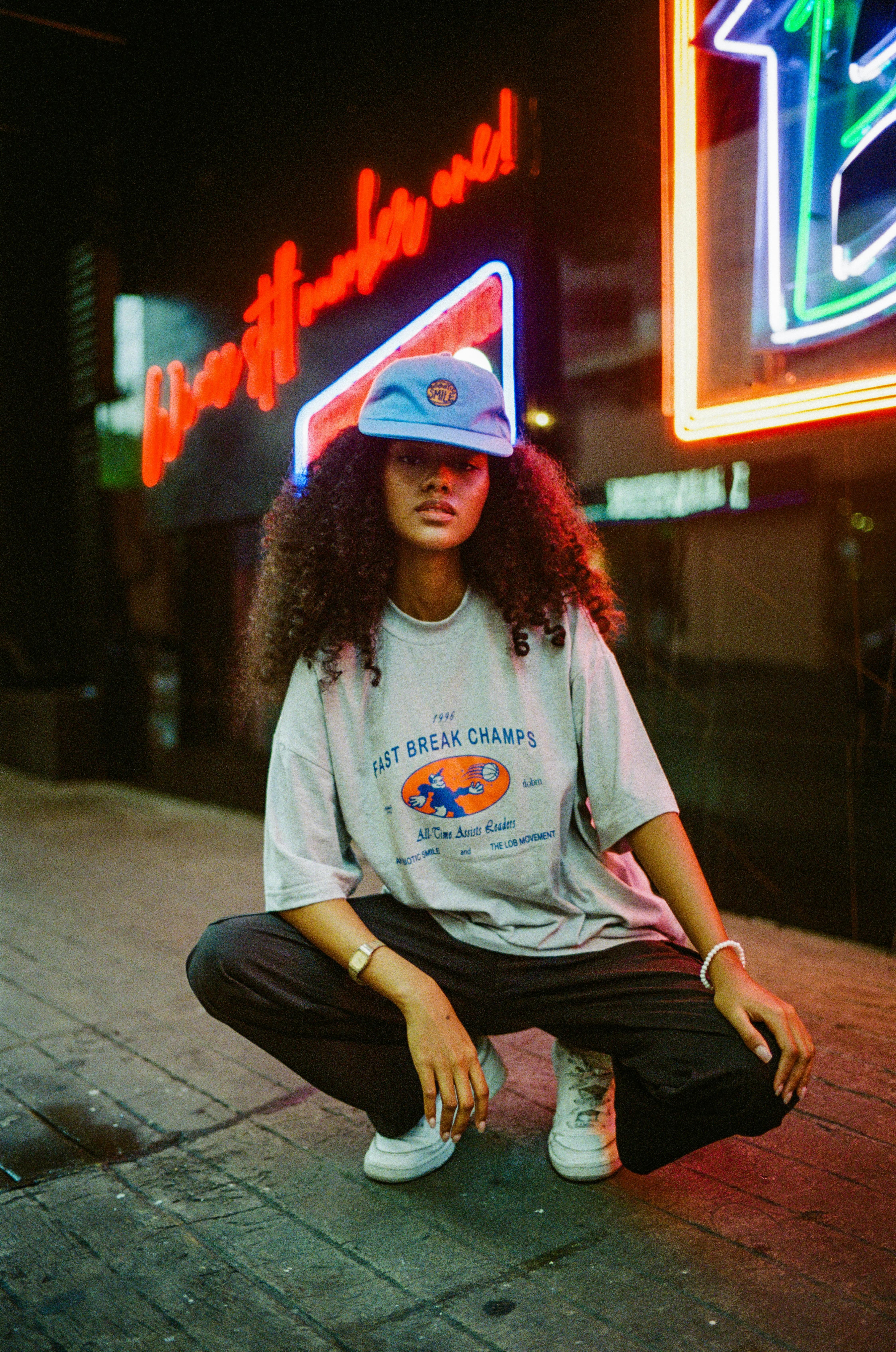 Young woman wearing an oversized graphic streetwear t-shirt, blue cap, and sneakers under neon city lights